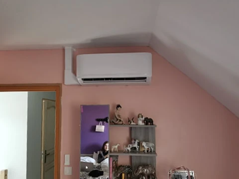 Chambre mansardée avec murs roses, climatiseur mural blanc installé près du plafond incliné